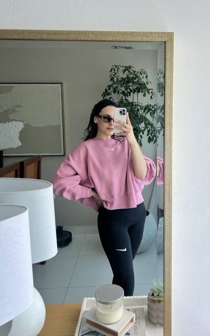 A woman takes a mirror selfie wearing a pink Nike sweatshirt and black leggings, holding a smartphone in a bright room with a lamp and plant visible.