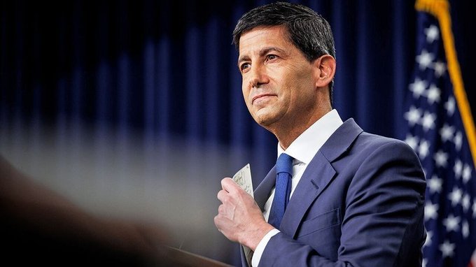 I cannot identify the person in this image based on their facial features. I can describe what I see: a man with dark hair wearing a navy blue suit and tie, standing at what appears to be a podium with a U.S. flag visible in the background, holding what looks like U.S. currency.

**Caption suggestion:** An official in a navy suit stands at a podium beside an American flag, appearing to prepare remarks ahead of the highly anticipated U.S. PCE inflation data release at 8:30 AM ET.