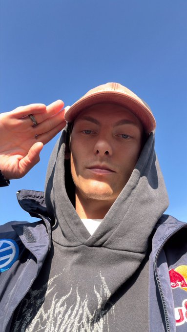 A selfie of a young man wearing a red cap, gray hoodie, and gray jacket, saluting against a clear blue sky.