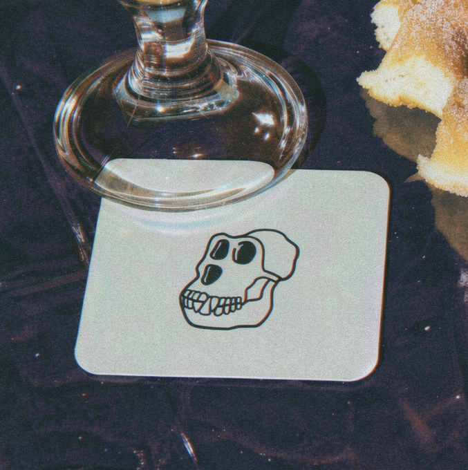 A cream-colored coaster featuring a black skull illustration sits beneath the base of a wine glass on a dark surface, paired with what appears to be a sugared pastry on the side—evoking a "happy hour" theme that matches the caption's quip about it always being 5 o'clock somewhere.