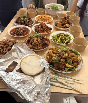A wooden table is set with a feast of various Chinese dishes in bowls and plates, accompanied by flatbread wrapped in foil and chopsticks, set for a group meal.