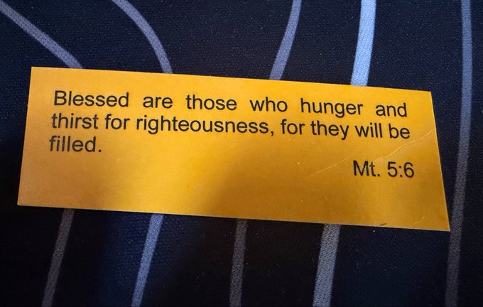 A yellow card resting on a striped fabric displays the text: 'Blessed are those who hunger and thirst for righteousness, for they will be filled. Mt. 5:6'.