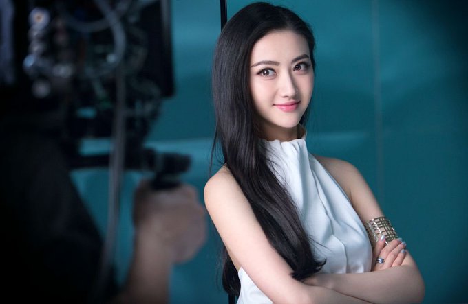 A young woman with long dark hair, wearing a white sleeveless top and a gold cuff bracelet, poses confidently with her arms crossed in front of a camera on a film set. The image relates to the post by humorously suggesting the woman could be casting for "Crypto Diggers," playfully drawing a parallel between her poised, glamorous appearance and the allure of gold-seeking prospectors now chasing cryptocurrency fortunes.