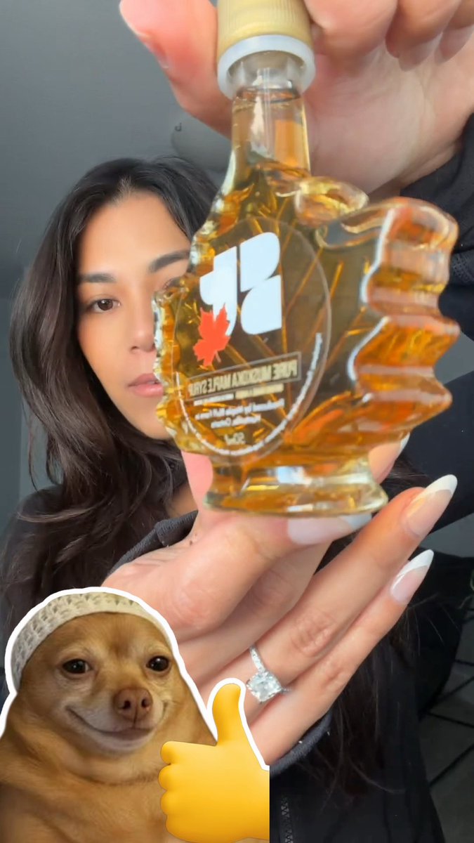 A woman holding up a maple syrup-shaped bottle labeled 'PURE MAPLE SYRUP' with a Canadian flag logo, accompanied by a sticker of a smiling dog giving a thumbs-up in the corner.