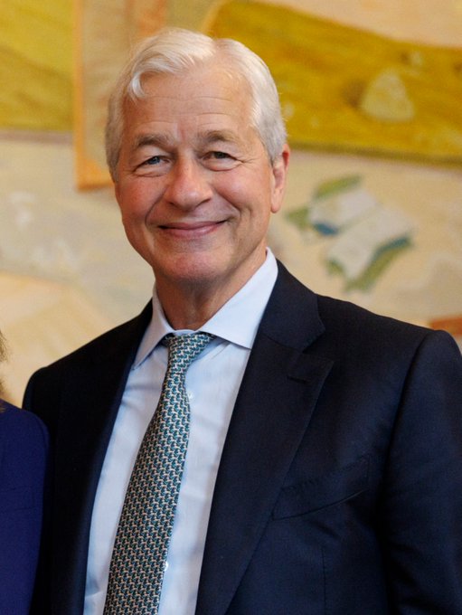 A portrait of a smiling older man with white hair wearing a dark navy suit, light blue shirt, and a green and blue patterned tie, standing in front of what appears to be a colorful painting.