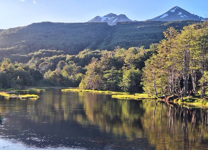 Scenic landscape photograph of a calm lake surrounded by lush green trees with snow-capped mountains in the background under a partly cloudy sky.