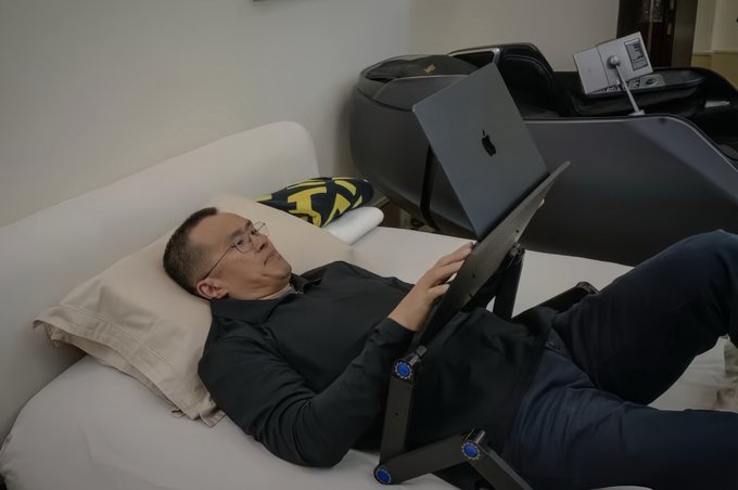 A man wearing glasses reclines on a white couch, working on a laptop with the Apple logo visible, while another Apple laptop and iPad sit on a black massage chair in the background, capturing a candid moment of CZ's everyday lifestyle that the poster idolizes.