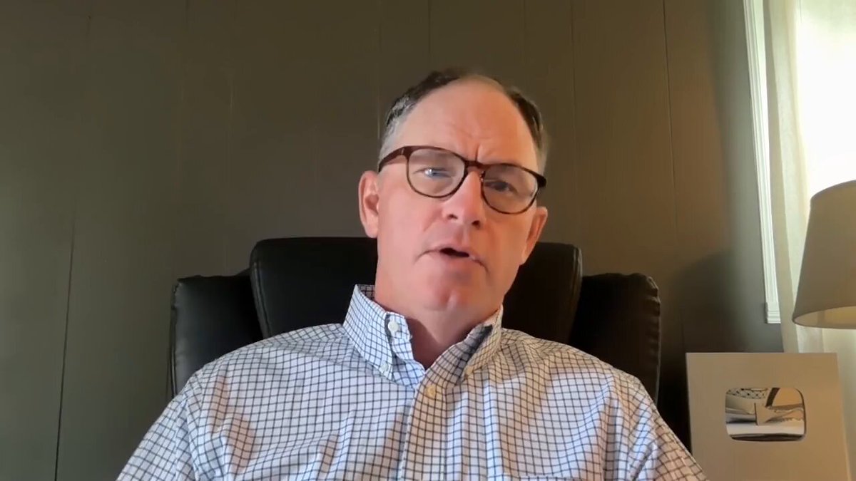 The image shows a man wearing glasses sitting in an office chair, with his head resting against a wall. He appears to be looking off into the distance or engaged in a conversation. The background features a window that allows natural light to enter the room. There is also a lamp on the desk next to him.