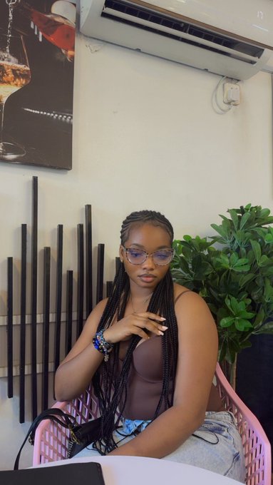 A young woman with long braided hair and glasses sits in a pink chair, posing indoors with a potted plant and decorative wall art behind her.