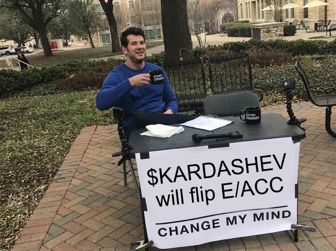 A man sitting outdoors at a 'Change My Mind' table holding a 'Steven Crowder' mug, with a sign reading '$KARDASHEV will flip E/ACC'.