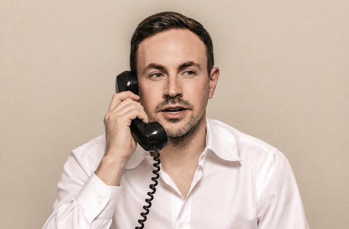 A studio portrait of a man in a white dress shirt holding a black vintage-style telephone receiver to his ear, looking off to the side against a beige background.