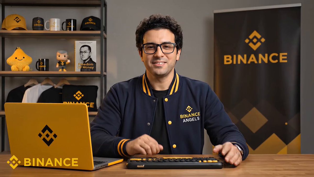 The image features a man sitting at a desk, wearing glasses and a blue jacket with "Binance" written across his chest. He is using a laptop computer placed next to him while smiling for the camera. The background of the image shows a poster or banner that says "Binance", indicating that this scene takes place in an environment related to Binance trading or stock market analysis.