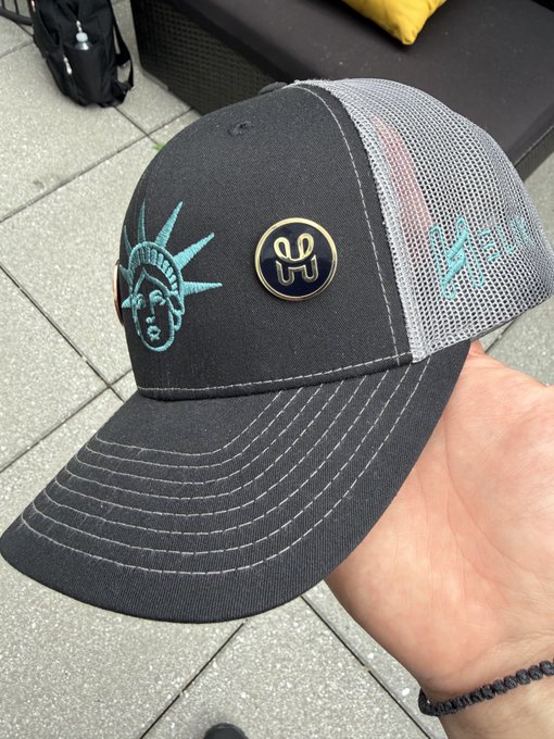 A hand holds up a black trucker hat featuring a Statue of Liberty design and a gold '000' logo pin, with a Liberty-themed graphic on the mesh side.