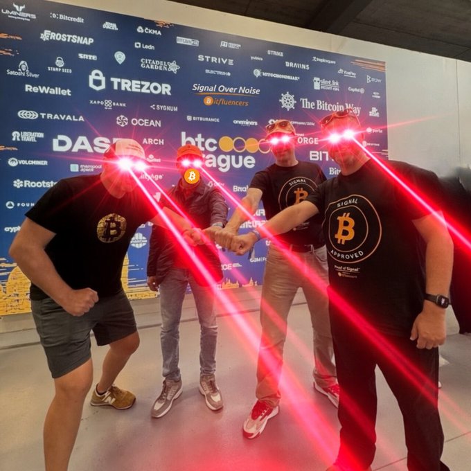 Four men pose with fists together in front of a backdrop featuring Bitcoin and crypto sponsor logos (including TREZOR, BTC Prague, Binance, Strive, Signal Over Noise, and The Bitcoin Way), wearing shirts branded with the "Bitcoin Signal Approved" logo, visually reinforcing the post's message of choosing Bitcoin-focused "signal" over AI-generated noise.