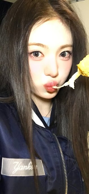 A close-up selfie shows a young woman with long dark hair making a pouting expression while holding a piece of fried food near her mouth; she wears a navy jacket with 'Yankees' text.
