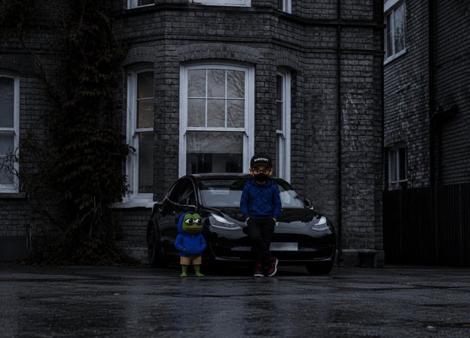 A child in a blue jacket stands beside a small green plush figure in front of a black Tesla parked on a wet street in front of a dark brick building.