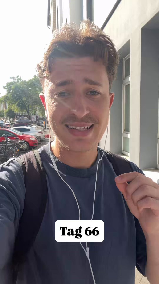 The image features a man wearing headphones, with his hand raised to his face as if he's about to make a statement or ask a question. The background includes several cars parked along the street, suggesting that this scene takes place in an urban setting. The caption of the image reads "Tag 66 bis Bitcoin auf 1 Million geht", which is likely related to the man and the post on social media platforms.