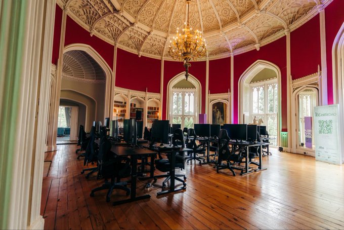 An ornate room with red walls, gothic arches, a chandelier, and wooden floors contains rows of computer desks and monitors.