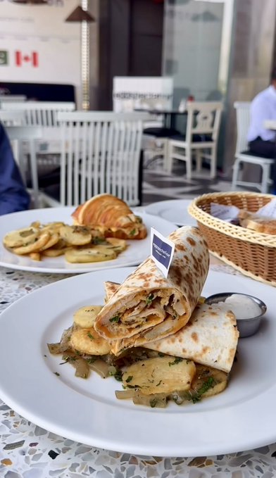 A shawarma wrap and a croissant with potatoes are served on white plates at a restaurant patio, with a small sign reading "SHAWARMA & CO" visible behind the dish, accompanying a post celebrating a 0.5 $SOL meal purchase.