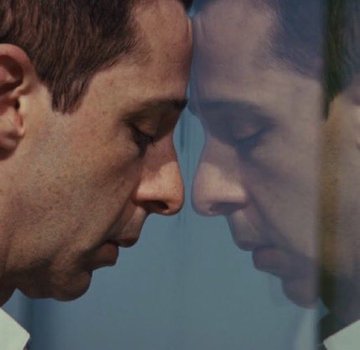 A close-up profile shot of a man pressing his forehead against a mirror, creating a mirrored reflection of his face.