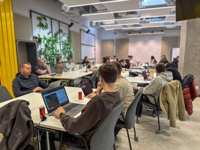 A group of people are working on laptops at a long table in a modern office or classroom setting with yellow curtains and indoor plants.