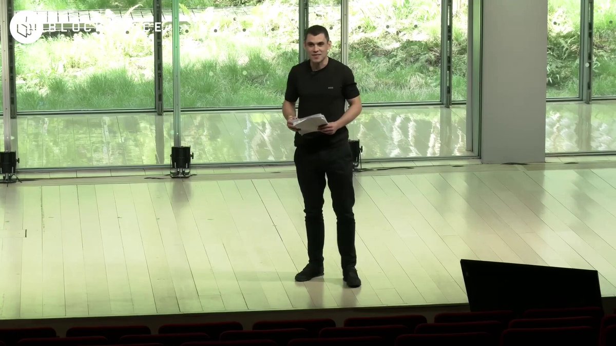 The image shows a man standing on stage, holding a piece of paper or a book while giving a speech. He appears to be addressing an audience, possibly at a conference or event. The setting includes a large window behind him that allows natural light into the room and provides a view of trees outside. The speaker's attire consists of a black shirt and pants, which contrasts with the white background of the stage.