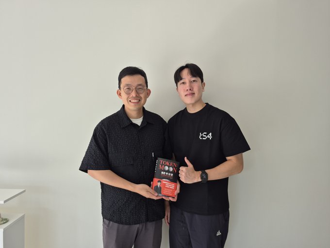 Two men pose together holding a red 'TOKEN MODEL' package; one wears glasses and a black shirt, the other wears a black 'tS4' t-shirt.