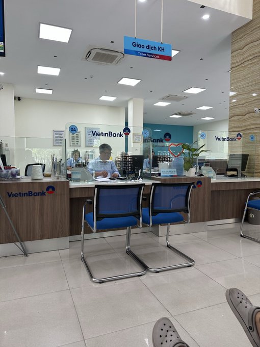 Interior of a VietinBank branch featuring teller stations numbered 04-05, with a 'Giao dịch KH / Teller Services' sign overhead and blue customer chairs in the waiting area.