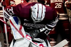 A Massachusetts hockey player wearing a maroon jersey and helmet sits in the locker room, leaning forward with their stick across their lap.
