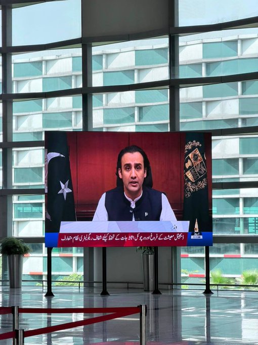 A large screen displays a man in a black vest addressing viewers, flanked by the Pakistani flag, with Urdu text banners below reading 'اسپیشل ٹرانسفارمیشن ایجنسی کا کرپٹ لوگوں کو قانون کی گرفت میں لانے کیلئے بھرپور ایکشن لیا جائے گا، عاصمہ' and a 'STATE LIFE' logo.