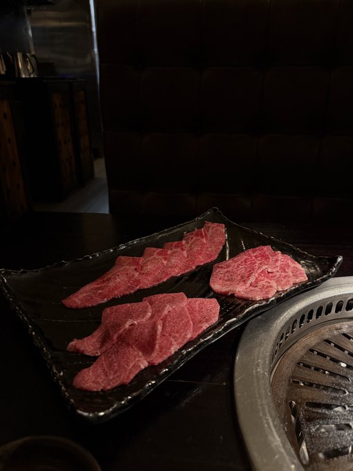 Three portions of raw, marbled meat presented on a textured black rectangular plate next to a circular metal grill in a dimly lit setting.