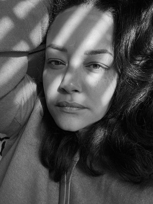 A black-and-white close-up portrait of a young woman with dark hair, illuminated by dramatic striped shadows across her face.