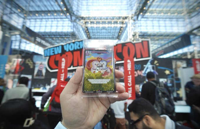 A hand holds up a foil trading card pack featuring a rainbow-colored character in front of a 'NEW YORK COMIC CON' banner inside a large convention hall.