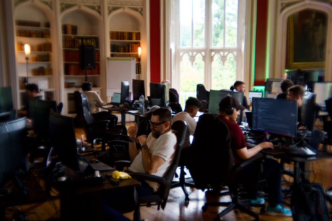 A group of developers works intently at multiple computer stations set up in an ornate, book-lined room with Gothic windows, blending modern tech work with classical architecture. The setting directly reflects the caption's reference to "devs shipping code inside a castle," showing programmers immersed in their screens within the grand, historic interior.