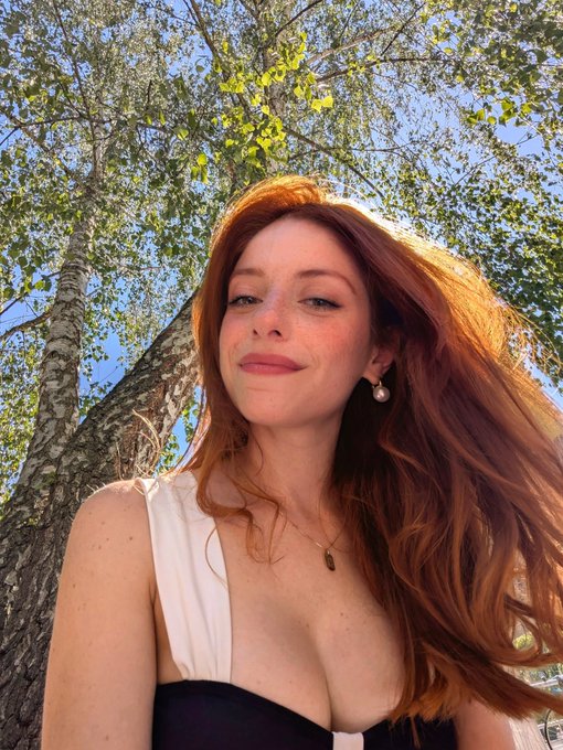 A young woman with long red hair smiles softly in an outdoor setting with green trees and blue sky behind her.