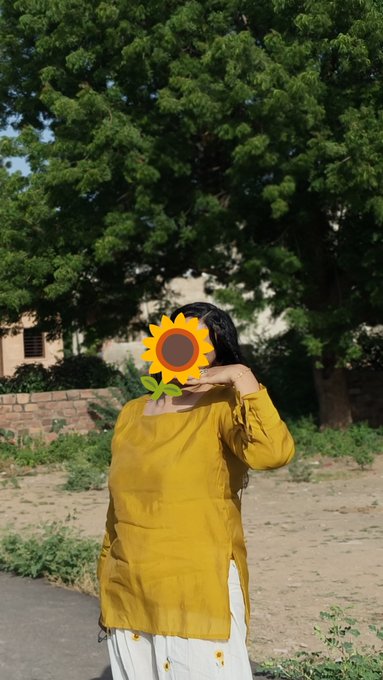 A photograph of a woman wearing a yellow tunic standing outdoors in front of a large tree, with a cartoon sunflower graphic covering her face.