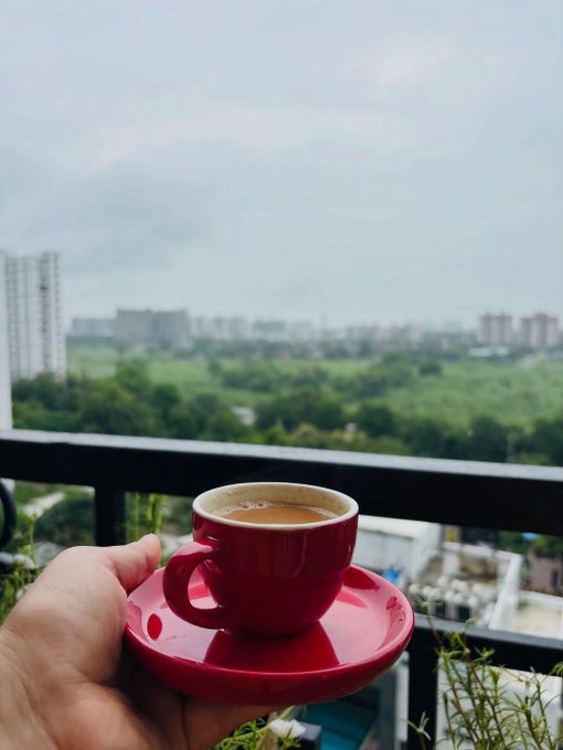 A hand holds a red cup of tea or coffee on a balcony overlooking a cityscape with green trees in the background.