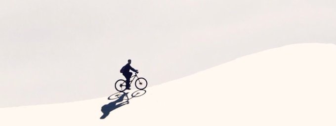 A minimalist illustration of a silhouette of a person riding a bicycle up a hill against a pale sky, casting a shadow on the ground.