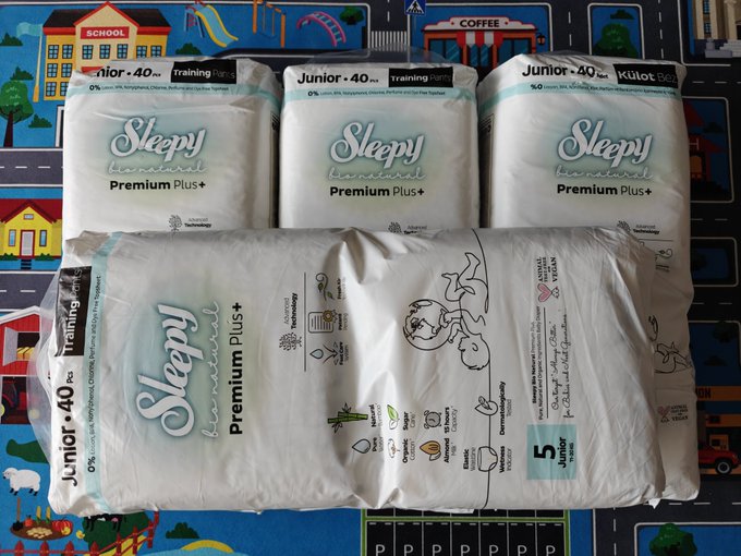 Four packages of "Sleepy brand Premium Plus+" junior diapers (size 40) are displayed on a colorful children's play mat featuring illustrations of a school, playground, and coffee shop, visually illustrating the post's humorous comparison of stockpiling diapers as a long-term "investment strategy" akin to dollar-cost averaging cryptocurrencies.