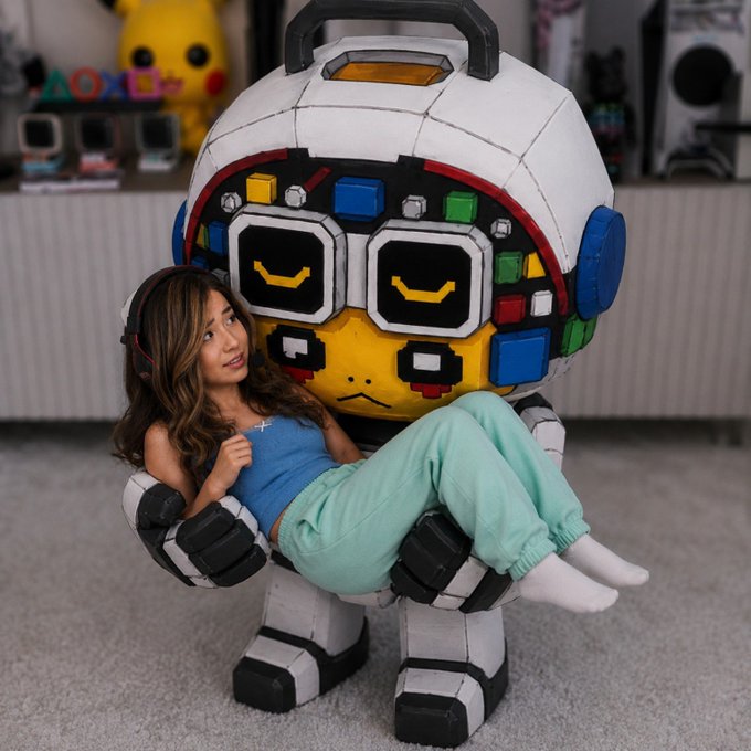 A young woman wearing headphones, a blue top, and mint green pants sitting in the arms of a large papercraft robot figure, in a room with gaming-related decor.
