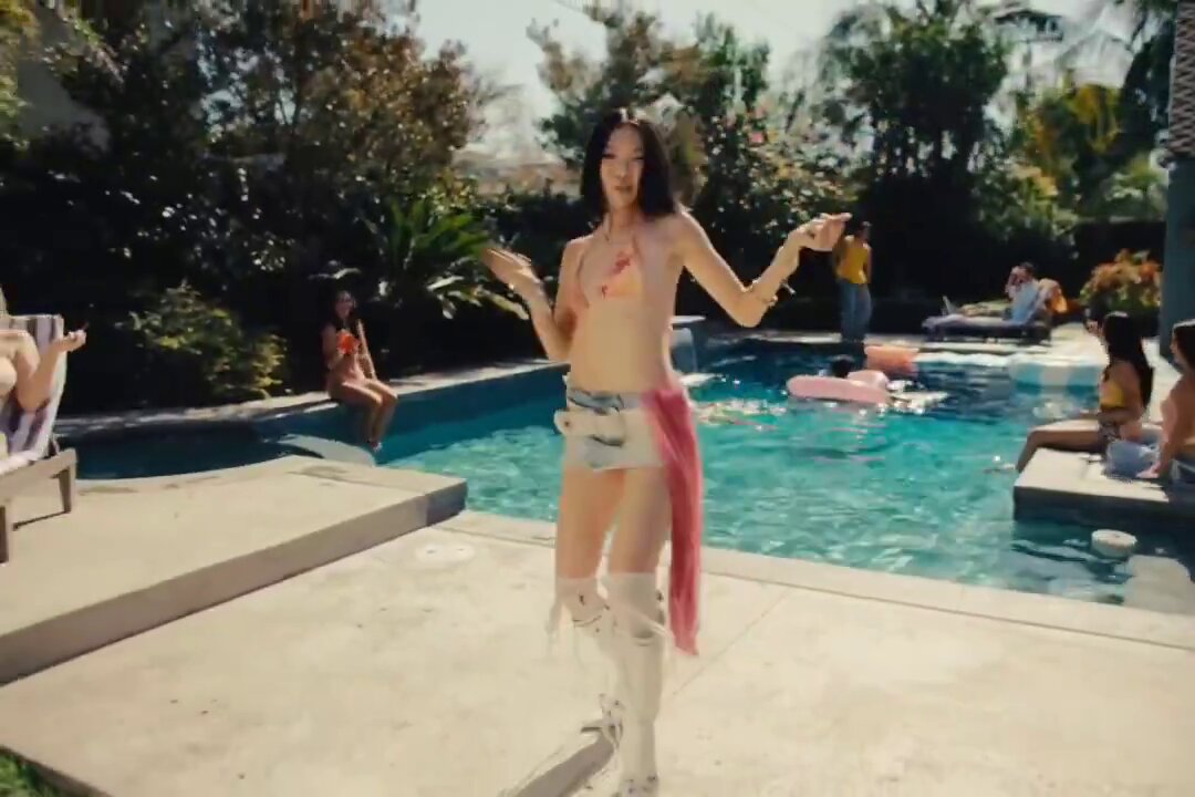 The image features a woman wearing a bikini top, shorts, and white boots standing by a pool at an outdoor party with several people around her. The photo captures the moment when she poses for the camera while holding her arms out wide. In the background, there are other people enjoying themselves in the pool area as well.