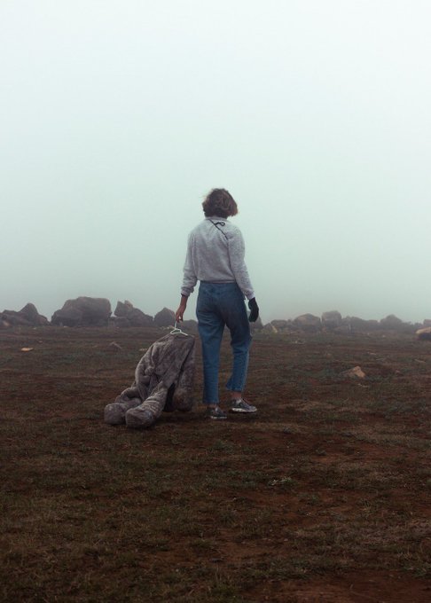 A person stands in a foggy, rocky landscape holding a stuffed teddy bear by its arm, captured in a moody, desaturated photographic style. The melancholic atmosphere and fine art aesthetic align with the minimalist, contemplative tone often featured by Masiha's photography on @mallowdotart.