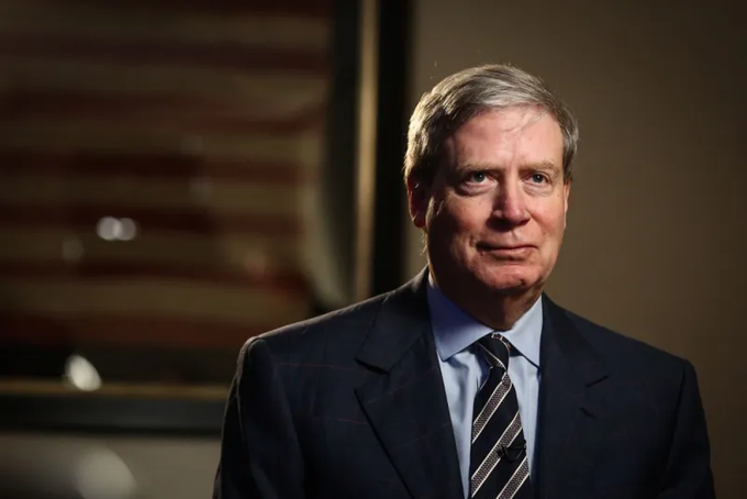 A portrait photograph of a middle-aged man with gray hair wearing a dark suit, light blue shirt, and striped tie, photographed in what appears to be an interview setting.