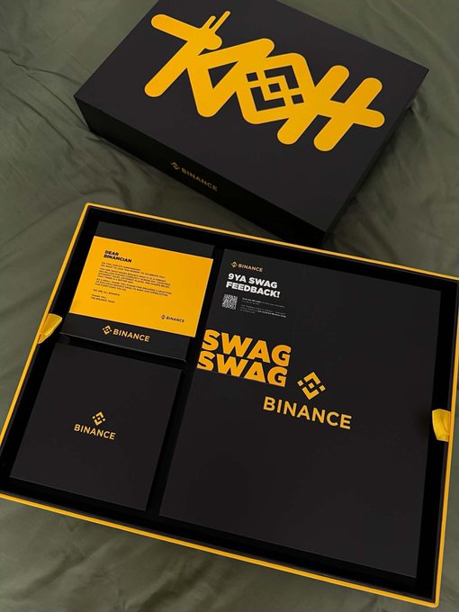 An open black and yellow Binance 'SYA SWAG FEEDBACK!' promotional box displaying branded merchandise and materials on a dark green surface.