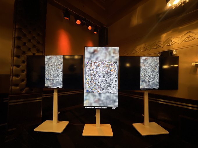 Three illuminated vertical screens display abstract, densely packed mixed-media artwork in a dimly lit gallery space, with the center screen labeled 'KAVINAS HERNANDES'.