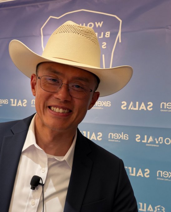 A smiling man wearing glasses, a cream-colored cowboy hat, white shirt, and dark blazer with a lavalier microphone, photographed in front of a backdrop with "U.S. SALES" branding.