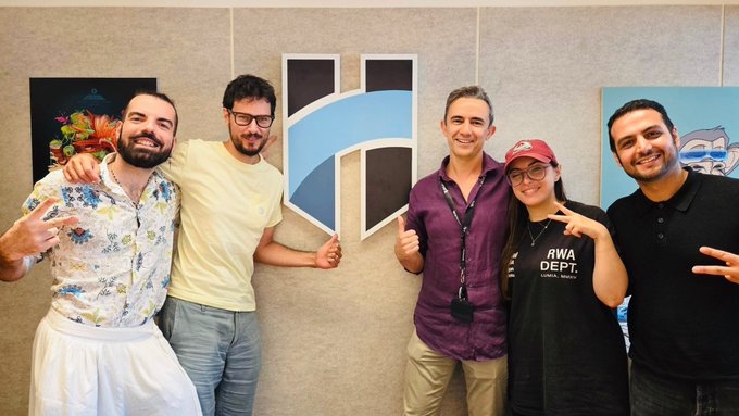 A group photo of five people posing together in front of a wall with an 'H' logo, including a woman wearing an 'AWA DEPT.' t-shirt and red cap, with team members smiling and gesturing.
