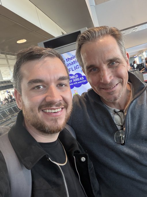 A selfie of two smiling men in an airport terminal, one younger with a beard and the other older with glasses hanging from his jacket, with a digital sign about flight areas visible behind them.