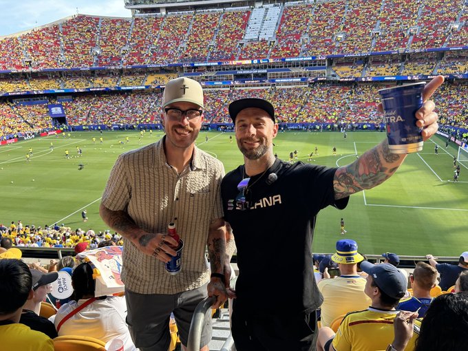 The image shows two men standing next to each other with their arms raised high above their heads, holding cups of beer as they pose for a picture. The background reveals that they are at a soccer stadium filled with fans watching the game. There is also a crowd of people in the foreground and behind them, indicating a lively atmosphere. The post accompanying this image reads: "I'm thankful, this is Solana Made Simple @solanamidwest at #FIFAWorldCup".