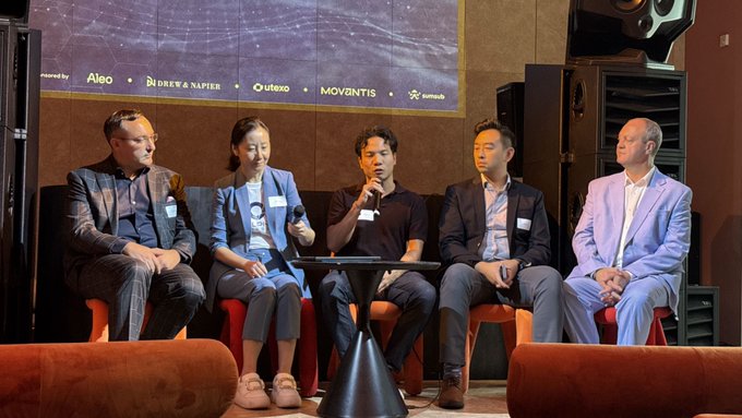 Five people seated on a stage during a panel discussion, with sponsor logos including Aleo, Durey & Napier, Utexo, and Movgntis visible on the screen behind them.